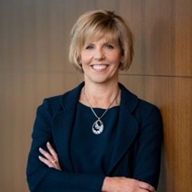 Professional woman with short blonde hair, wearing a dark blazer and a necklace, stands confidently with arms crossed against a wooden background.