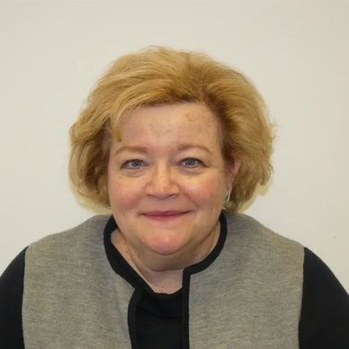 Smiling woman with short, curly blonde hair, wearing a gray cardigan over a black shirt, against a plain white background.