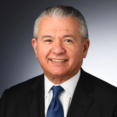 Professional headshot of a middle-aged man in a dark suit and tie, smiling against a gradient gray background. Suitable for business or networking contexts.