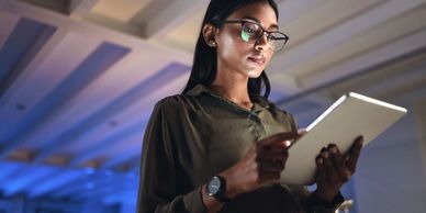 Woman with glasses focused on a tablet, illuminated by soft blue light in a modern workspace. Highlights remote work and digital engagement.